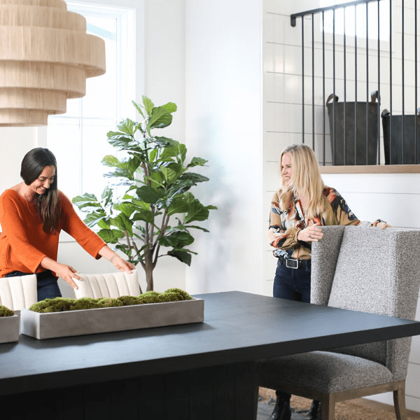 Women arranging furniture and decor in room.