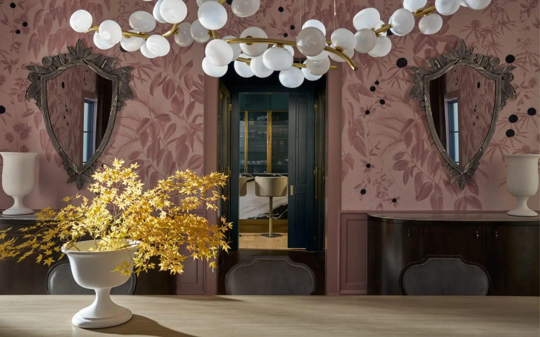 Sophisticated dining room with pink floral wallpaper, mauve wall paneling, branch-like chandelier and yellow leaves on the table.